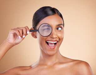 Face, beauty or magnifying glass with a model woman in studio on beige background searching for skincare. Facial, eye wink or excited Indian girl looking for luxury self care cosmetics or makeup