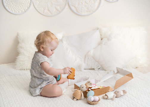 A girl in a white dress on her head plays on the bed with a box of gingerbread in the form of Easter eggs. Horizontal photo. Light background. Family and home holiday concept. High quality photo