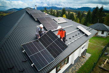 Engineers building photovoltaic solar module station on roof of house. Men electricians in helmets installing solar panel system outdoors. Concept of alternative and renewable energy. Aerial view. - obrazy, fototapety, plakaty