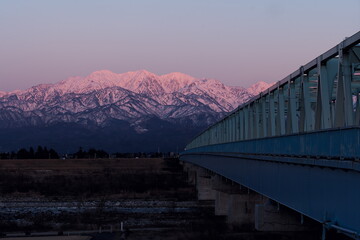 常願寺川に架るダイナミックな道路鉄橋と夕焼けに染まる冠雪の北アルプス, 毛勝三山, 毛勝山