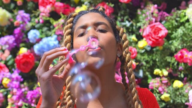 Beautiful young latino lady with cool hair braids blowing soap bubbles with colorful flowers background - dreamy aesthetic concept