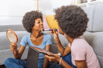 Black sisters, one applying make-up to the other, sitting on the living room floor