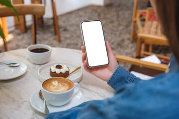 Mockup image of a woman holding and using mobile phone with blank desktop screen with cake and coffee cups on the table in cafe