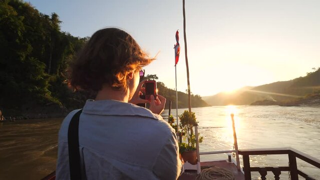 Young Tourist Woman Takes Pictures of Beautiful Sunset at Mekong River Cruise Trip on Wooden Slow Boat from Luang Prabang, High Quality 4K Slowmotion Tourism in South East Asia Concept Footage, Laos.