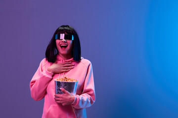 Happy excited pretty brunet woman in pink hoodie trendy specular sunglasses with popcorn hold hand on chest posing isolated in blue violet color light background. Neon party Cinema concept. Copy space