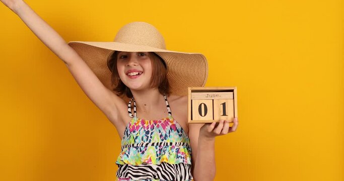 Child teenager girl in swimsuit and straw hat hold in hand wooden calendar on 01 June in studio on yellow background, summer start, summer mood, World Children's Day