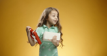 Cute little girl opening her gift, surprised by the contents, looking around and happily smiling, isolated on yellow background - emotions concept. Close up. Copy space