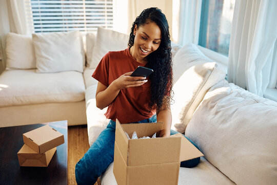 Happy black woman using cell phone while unpacking delivery box at home.