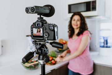 Latin woman influencer recording video class or live stream with camera about healthy eating and nutrition in kitchen at home in Mexico, hispanic people