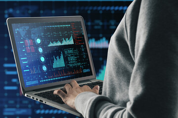 Close up of hacker hand using laptop with glowing digital interface with business charts and coding on dark background. Future and cyberspace concept.