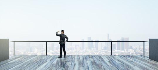 Back view of young businessman looking into the distance on rooftop with bright daylight sky and wide city view. Success, future and vision concept.