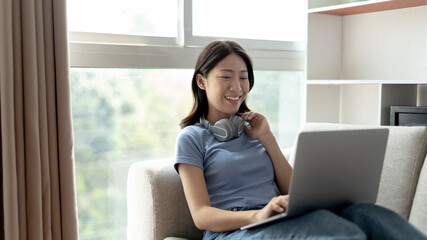 Asian woman sitting on the sofa with a laptop, Looking at laptop screen, Relaxing at home, Working through the Internet communication system, Happy lifestyle, Feel good.