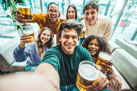 Happy friends taking selfie picture at brewery pub restaurant - Group of multiracial people enjoying happy hour drinking beer sitting at bar table - Life style concept with guys and girls hanging out