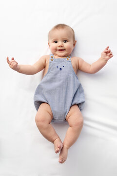 Smiling baby boy lying on white bed wearing blue checked overalls