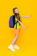 A little girl with a school backpack. A child in summer shorts is going to educational courses.  A happy child with a satchel. A full-length schoolgirl on a yellow isolated background.