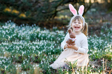 Happy little girl with Easter bunny ears eating chocolate figure in spring forest on sunny day,...