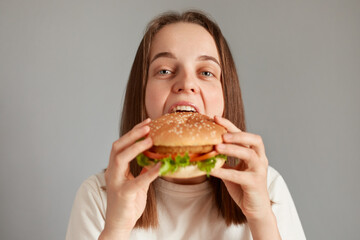 Indoor shot of hungry woman with brown hair biting cheeseburger with pleasure, eating hamburger,...