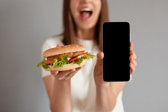 Image of unrecognizable unknown woman wearing white t-shirt holding fast food burger and showing mobile cell phone with blank screen isolated on gray background.