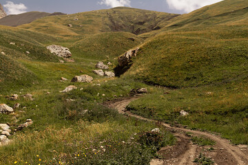 Obraz premium Mountain landscape - hilly mountain valley with country road and big boulders in fine sunny summer day, green and yellow velvety meadow on with slopes. Awe travel in Dagestan.