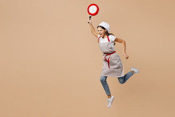 Full body sideways mad sad young housewife housekeeper chef baker latin woman wear apron toque hat run fast threat with frying pan isolated on plain pastel light beige background. Cook food concept.