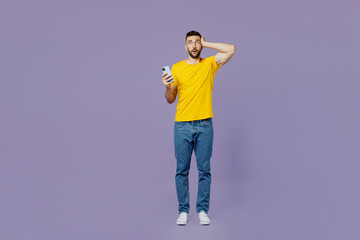 Fototapeta premium Full body young sad shocked fearful caucasian man wear yellow t-shirt use mobile cell phone hold head look camera isolated on plain pastel light purple background studio portrait. Lifestyle concept.