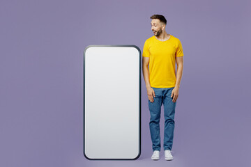 Full body young surprised shocked amazed impressed fun man wear yellow t-shirt looking at big huge blank screen mobile cell phone smartphone with area isolated on plain pastel light purple background.