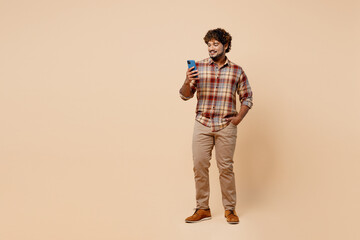 Fototapeta premium Full body smiling happy young Indian man wear brown shirt casual clothes hold in hand use mobile cell phone isolated on plain pastel light beige background studio portrait People lifestyle concept.