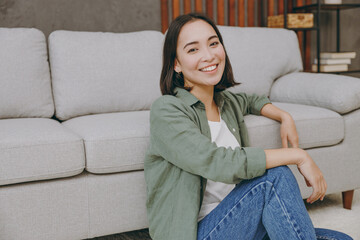 Obraz premium Young smiling happy cool fun satisfied woman of Asian ethnicity wears casual clothes look aside sit near grey sofa couch stay at home hotel flat rest relax spend free spare time in living room indoor
