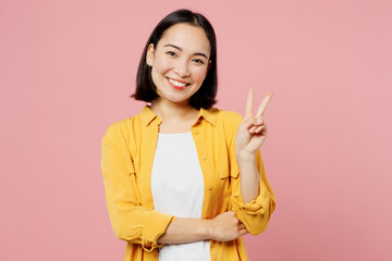 Young smiling happy cheerful friendly woman of Asian ethnicity wear yellow shirt white t-shirt showing victory sign isolated on plain pastel light pink background studio portrait. Lifestyle concept.