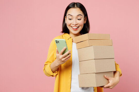 Young surprised happy woman of Asian ethnicity wears yellow shirt white t-shirt hold stack cardboard blank boxes use mobile cell phone isolated on plain pastel light pink background studio portrait.