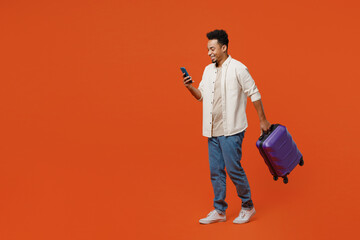 Full body traveler man wears casual clothes hold suitcase use mobile cell phone isolated on plain plain orange red background. Tourist travel abroad in free spare time rest. Air flight trip concept. - obrazy, fototapety, plakaty