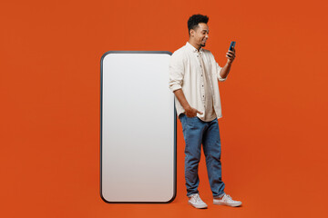 Full body young man wear light shirt casual clothes big huge blank screen mobile cell phone smartphone with copy space mockup area hold in hand use mobile cell phone isolated on orange red background.