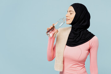 Young thirsty arabian asian muslim fitness trainer sporty woman wear pink abaya hijab towel spend time in home gym drink water isolated on plain blue background studio. Workout sport fit abs concept.