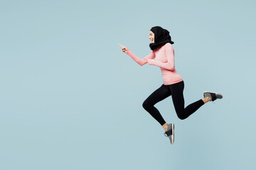Full body young asian muslim fitness trainer sporty woman wear pink abaya hijab spend time in home gym jump high point aside run isolated on plain blue background studio Workout sport fit abs concept