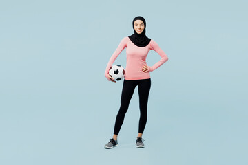Fototapeta na wymiar Full body smiling young asian muslim fitness trainer sporty woman wear pink abaya hijab spend time in home gym hold soccer ball isolated on plain blue background studio. Workout sport fit abs concept.