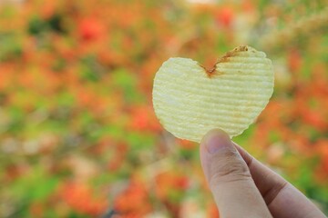 Heart-shaped potato chip in woman hand with colorful leaves in autumn background. Food and Valentine's day concept.
