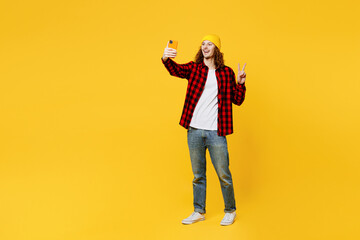 Fototapeta na wymiar Full body young man wearing red checkered shirt white t-shirt hat doing selfie shot on mobile cell phone post photo on social network show v-sign isolated on plain yellow background studio portrait.
