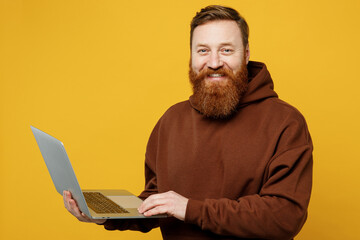 Side view smiling happy young redhead IT man wearing brown hoody casual clothes hold use work on laptop pc computer look camera isolated on plain yellow background studio portrait. Lifestyle concept.