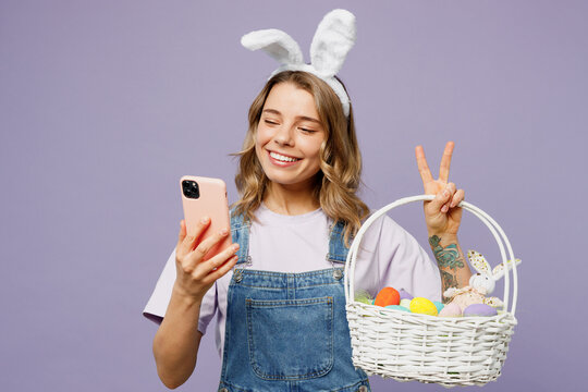Young fun woman wearing casual clothes bunny rabbit ears hold wicker basket colorful eggs use mobile cell phone show v-sign isolated on plain purple background studio portrait. Happy Easter concept.