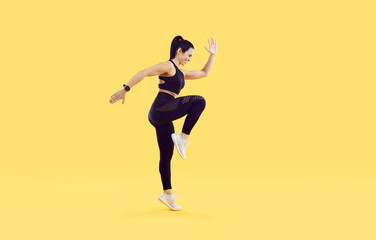 Fototapeta na wymiar Active sporty woman doing high knees exercise isolated on vivid yellow background. Full length side view of smiling woman with beautiful athletic body dressed in black sportswear doing sports workout.