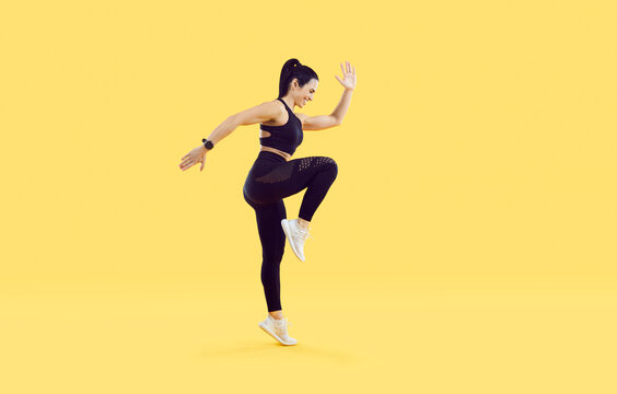Active sporty woman doing high knees exercise isolated on vivid yellow background. Full length side view of smiling woman with beautiful athletic body dressed in black sportswear doing sports workout.