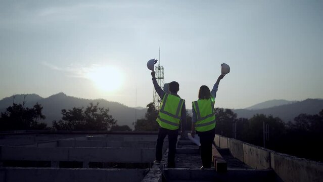 Silhouette of two engineers with helmets at construction site in background, sunset.