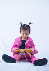 A portrait of a 2 year old girl sitting down, reading colored book on white isolated background. 