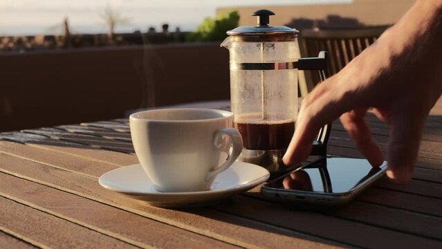 Caf&eacute; caliente humeante de cafetera de &eacute;mbolo sobre una mesa de madera con luz de amanecer en una terraza con vistas al mar y jardinera de fondo, con mano de hombre que coge un m&oacute;vil y unas gafas.
