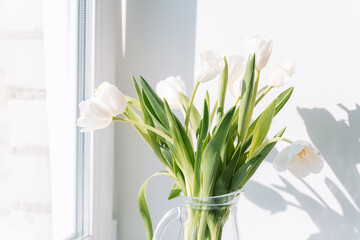 Bunch of white tulips in a glass decanter on the windowsill.