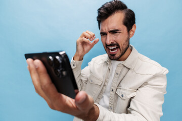 Portrait of a stylish brunette man looking at the phone blogger with a beard of anger and frustration, losing the game on a blue background in a white T-shirt and jeans, copy space