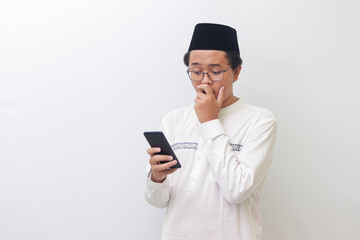 Portrait of young Asian muslim man holding mobile phone and covering mouth with shocked expression on face. Isolated image on white background