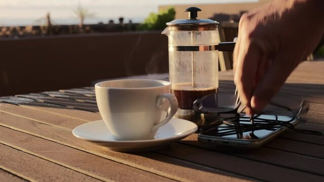 Caf&eacute; caliente humeante de cafetera de &eacute;mbolo sobre una mesa de madera con luz de amanecer en una terraza con vistas al mar y jardinera de fondo, con mano de hombre que coloca un m&oacute;vil y unas gafas.