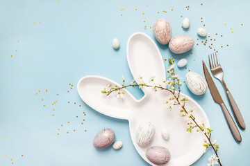 beautiful light easter mockup with bunny plate and cutlery, gold and marble eggs, cherry blossoms and confetti on a pastel blue background. top view. copy space. flat lay. place for text
