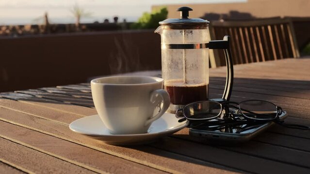 Caf&eacute; caliente humeante de cafetera de &eacute;mbolo sobre una mesa de madera con luz de amanecer en una terraza con vistas al mar y jardinera de fondo.